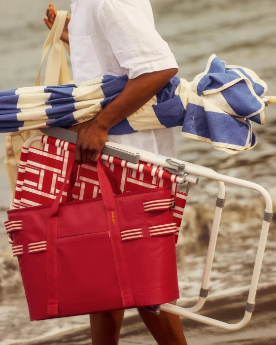 Person holding a red tote bag with striped towel, umbrella, and chair by water