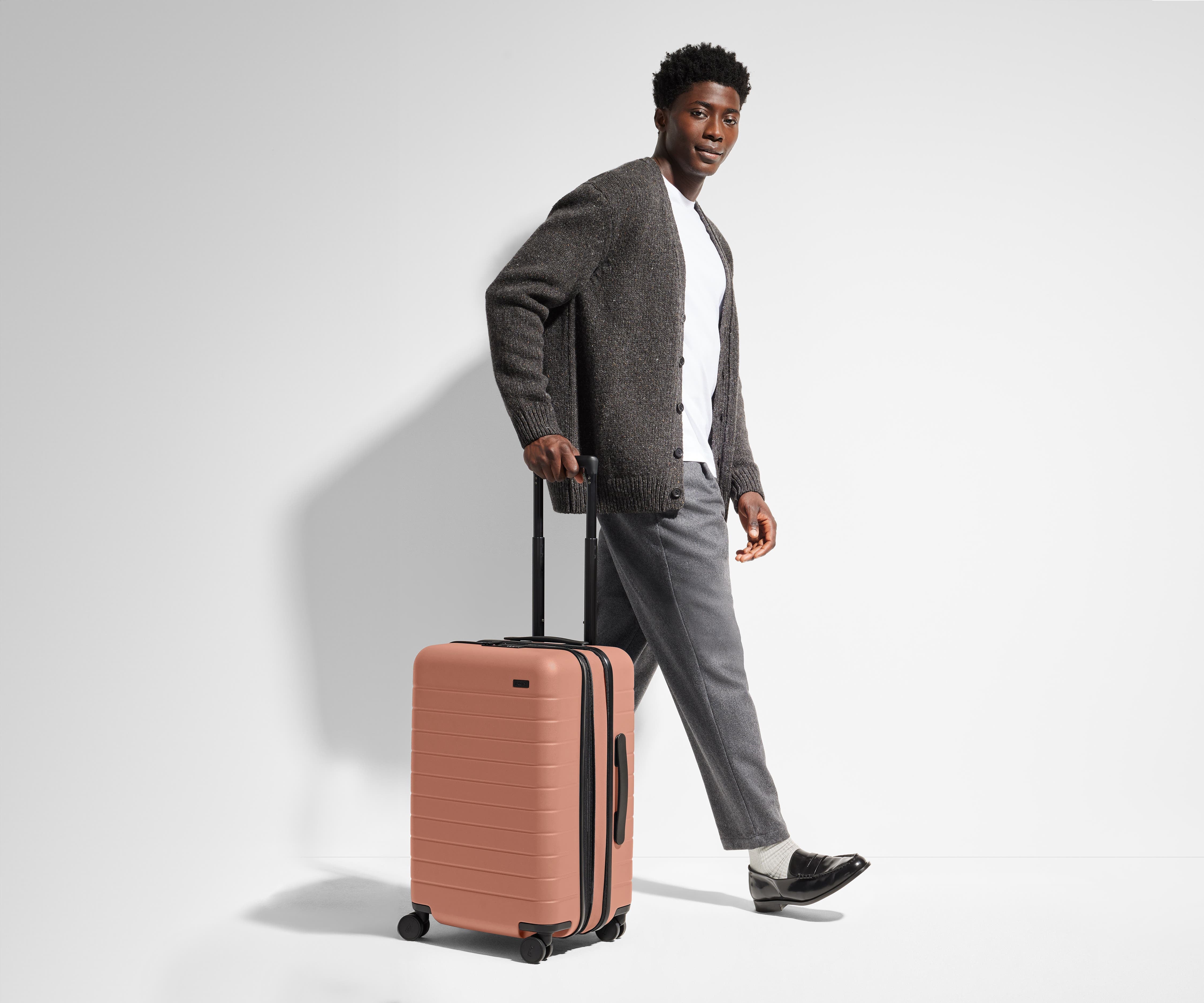 A 6'1" male model is standing next to and walking with The Bigger Carry-On suitcase by holding the raised handle to show size and scale