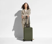 A 5'6" female model is standing next to The Bigger Carry-On suitcase and leaning her arms on the raised handle to show size and scale