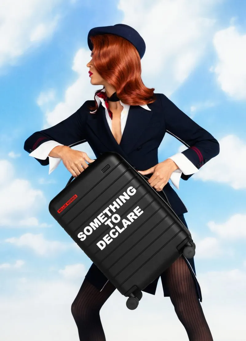 West Dakota holding an Away suitcase against a background with a cloudy, blue sky