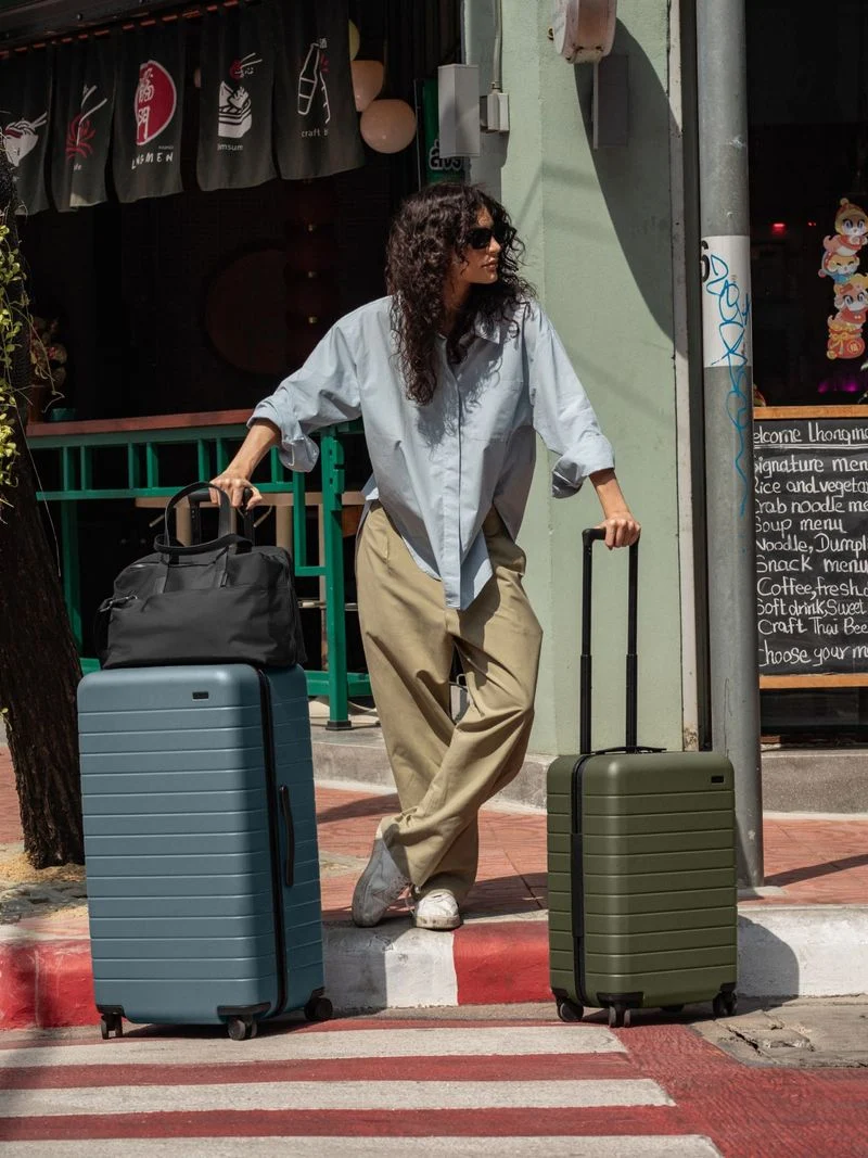 A woman in the streets of Bangkok with The Trunk in Coast Blue, The Carry-On in Olive Green, The Everywhere Bag in Black